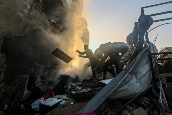 Palestinians search through the wreckage of buildings bombed by Israel in Khan Yunis, Gaza with their bare hands, with smoke in the background.