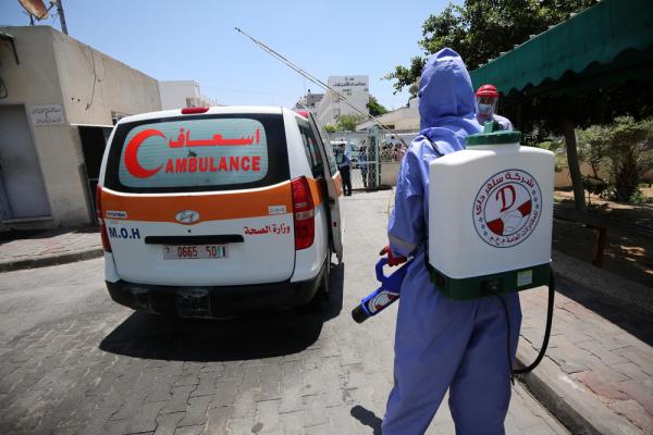 A worker in personal protective equipment carries a sanitation device and sprayer, walking near an ambulance during a COVID-19 preparedness exercise in Gaza City. July 2020.