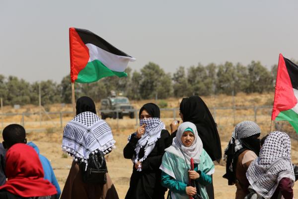 Palestinian girls protest at the Israel-Gaza border fence as part of the Great March of Return, some carry flags and/or wear kuffiyehs. On the other side of the fence, an Israeli mliitary jeep faces the girls. 2019.