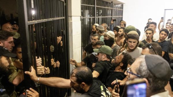 A large crowd of Israeli protestors storm the Beit Lid army base, with soldiers and police standing behind iron bars, attempting to keep protestors out.