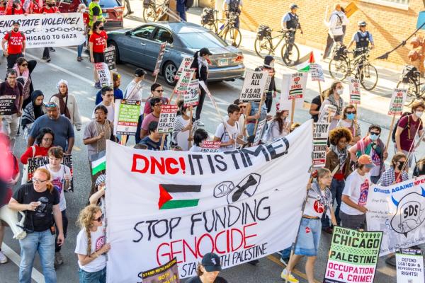 Protestors march on the DNC in Chicago, many carry signs demanding a ceasefire and expressing solidarity with Palestine. A large banner says "DNC, IT IS TIME! STOP FUNDING GENOCIDE."