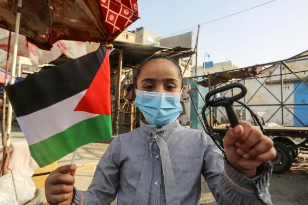 A Palestinian girl, carrying a Palestinian flag in one hand, and a key symbolizing the Palestinian right of return attends a Nakba commemoration event in Gaza. She wears a mask, as this event was held during the COVID-19 pandemic. May 2020.