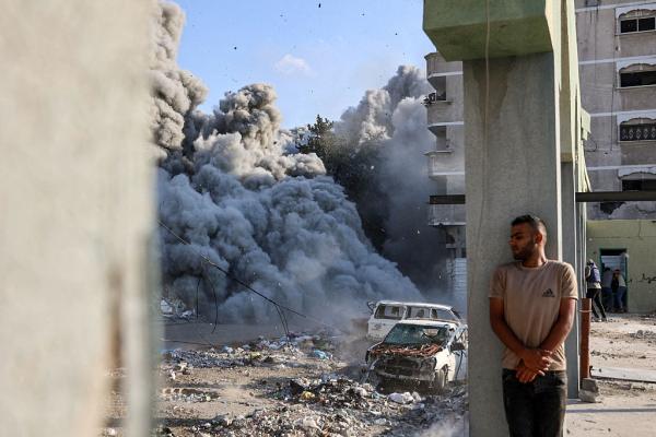 A Palestinian man takes cover from an Israeli bombing in the Zeitoun neighborhood of Gaza City, image shows large clouds of smoke and two vehicles destroyed by Israel's bombs.