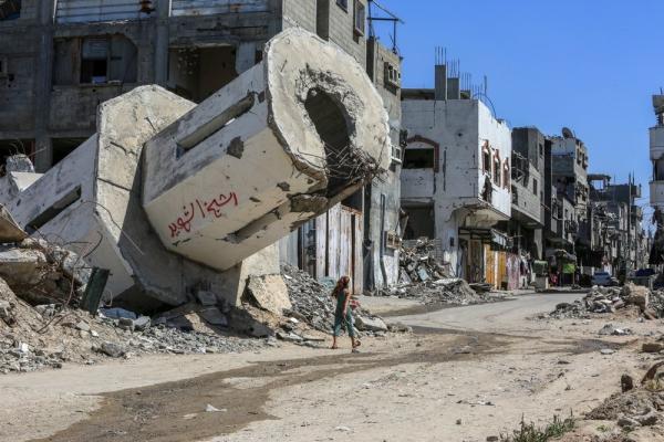 A Palestinian girl walks past a heavily damaged building pillar, hanging over her head, as she walks down a detroyed street in Gaza.