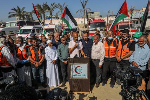 Aid workers, including health professionals and paramedics, participate in a press conference at the Nasser Medical Complex. PRCS ambulances are in the background, some participants hold Palestinian flags.