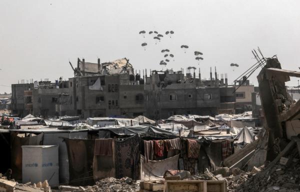 Aid aidrop falls over Rafah, with displacement tents and rubble in view.