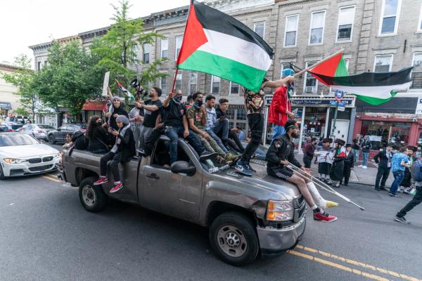 Arab and Muslim American youth protest Israel's 2021 war on Gaza in New York City, carrying Paelstinian flags and wearing kuffiyehs as they ride on the back of a pickup truck.