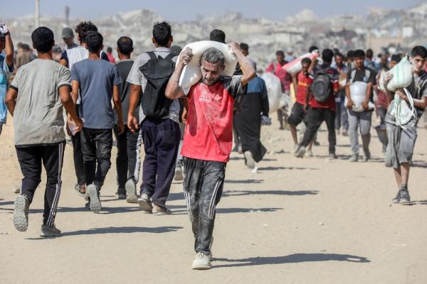 A Palestinian man walks with a large sack of flour he received, with flour on his body, as many others walk towards the aid distribution site.