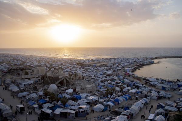 Sunset over a crowded makeshift displacement camp along the Gaza seaport, with destroyed rubble of a mosque minaret in view.
