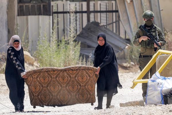 Palestinian women carry their sofa and a few personal belongings from their homes that they were expelled from in the Nur Shams camp near Tulkarm as an Israeli soldier watches. June 16, 2026.