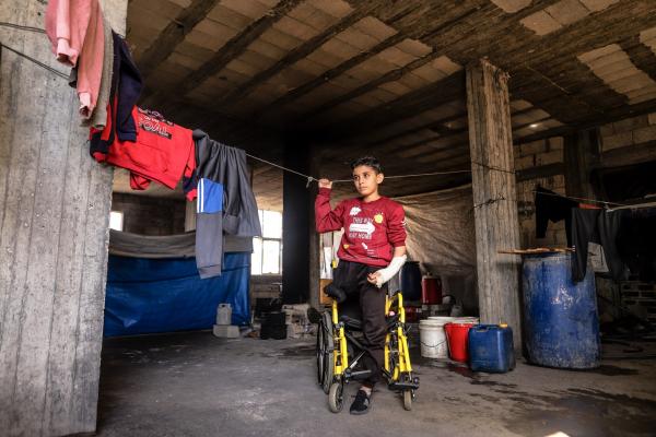 A 12-year-old boy stands on one leg in front of his wheelchair. His other leg was amputated due to an Israeli airstrike on Gaza.