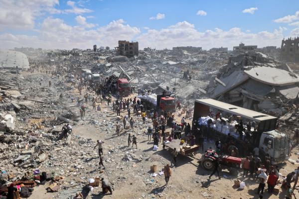 Palestinians wait for aid as trucks unload near the Kerem Shalom crossing in Khan Yunis on October 12, 2025. Aerial view shows total destruction of surrounding buildings and structures.
