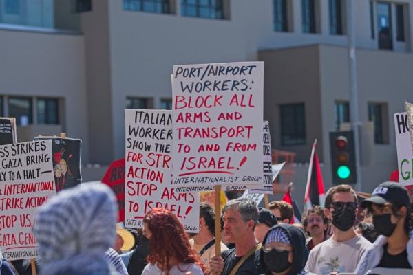 Protestors rally at a Rise Up for Gaza Event in Los Angeles, California. A protestor holds a sign that reads, "PORT/AIRPORT WORKERS: BLOCK ALL ARMS AND TRANSPORT TO AND FROM ISRAEL!"
