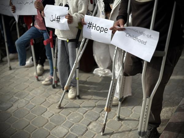Palestinian children who were forced to have limbs amputated due to Israel's genocidal war on Gaza appear at a rally in front of Nasser Hospital in Gaza. Image focuses on their crutches and amputated limbs, and the protest signs they carry, which read "genocide of childhood," and "hope."